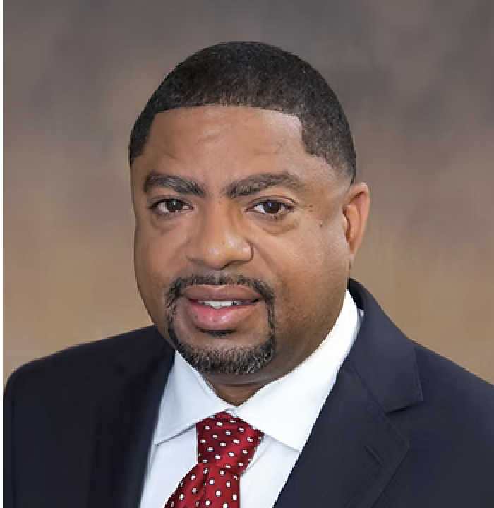 Professional headshot of a man in navy suit and red polka-dot tie against neutral background