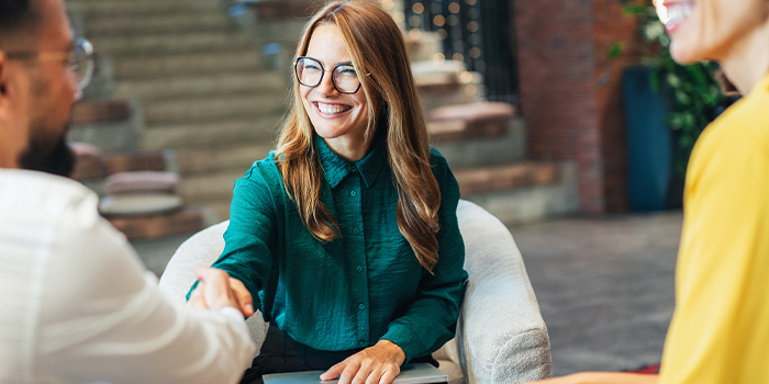 Smiling woman in green blouse and glasses shaking hands with client during casual office consultation