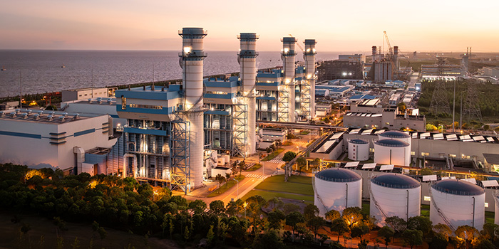 Coastal natural gas power plant at dusk with turbine stacks, storage tanks, industrial buildings and transmission lines.