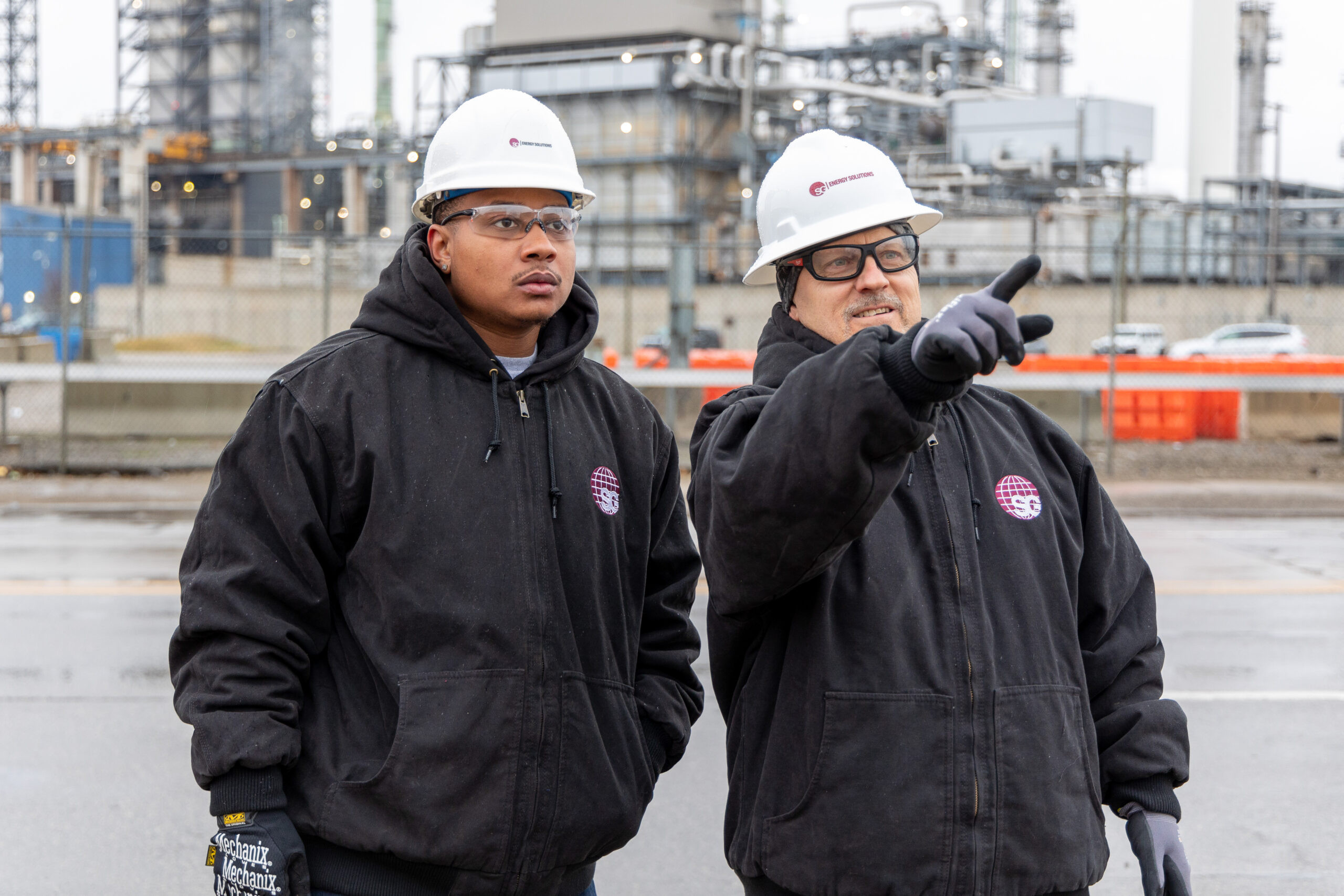 Two industrial workers wearing hard hats and jackets inspect a refinery plant; one points ahead.