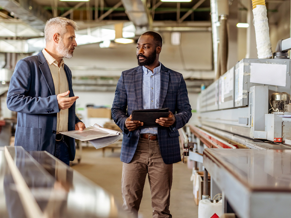 Two managers inspecting a production line in a factory, reviewing plans and a tablet during a facility walkthrough