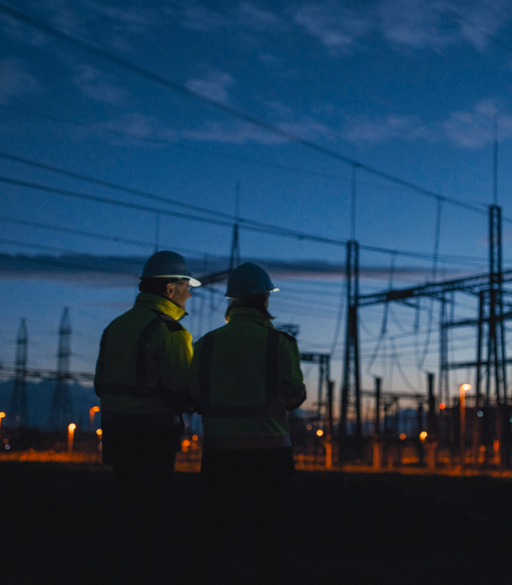 Two utility workers in hi-vis jackets and hard hats inspecting a power substation at dusk