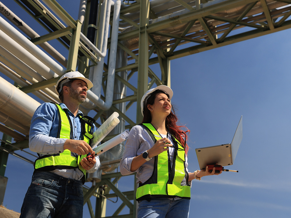 Two engineers in hard hats and hi-vis vests inspecting industrial plant, one with blueprints and one with laptop